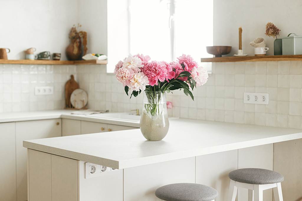 Beautiful peonies in vase on granite countertop island on background of stylish white kitchen with wooden shelves and appliances in new house. Modern minimal kitchen interior